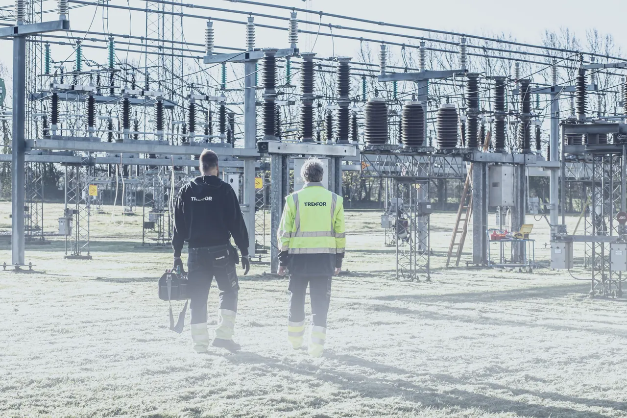 Two crew members walking through a substation yard, seen from behind.