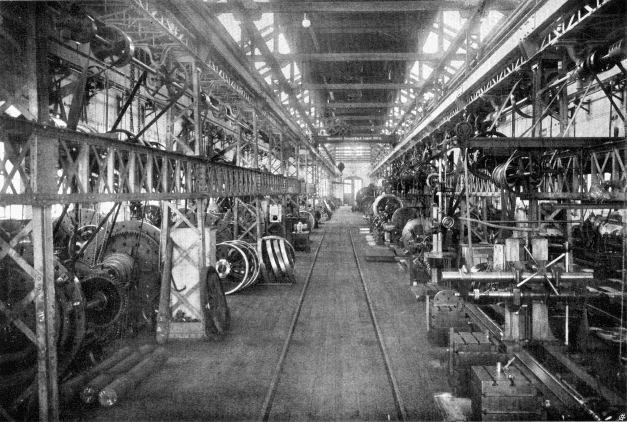 A long machine hall in 1910: line shafts run overhead the length of the building, with belts dropping to every lathe and wheel machine along a central track.